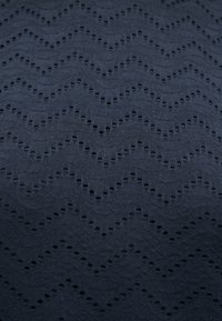 Σκούρο ύφασμα με υφασμένο ζιγκ-ζαγκ σχέδιο και ομοιόμορφα κατανεμημένες μικρές κυκλικές οπές που δημιουργούν ένα κυματιστό μοτίβο.