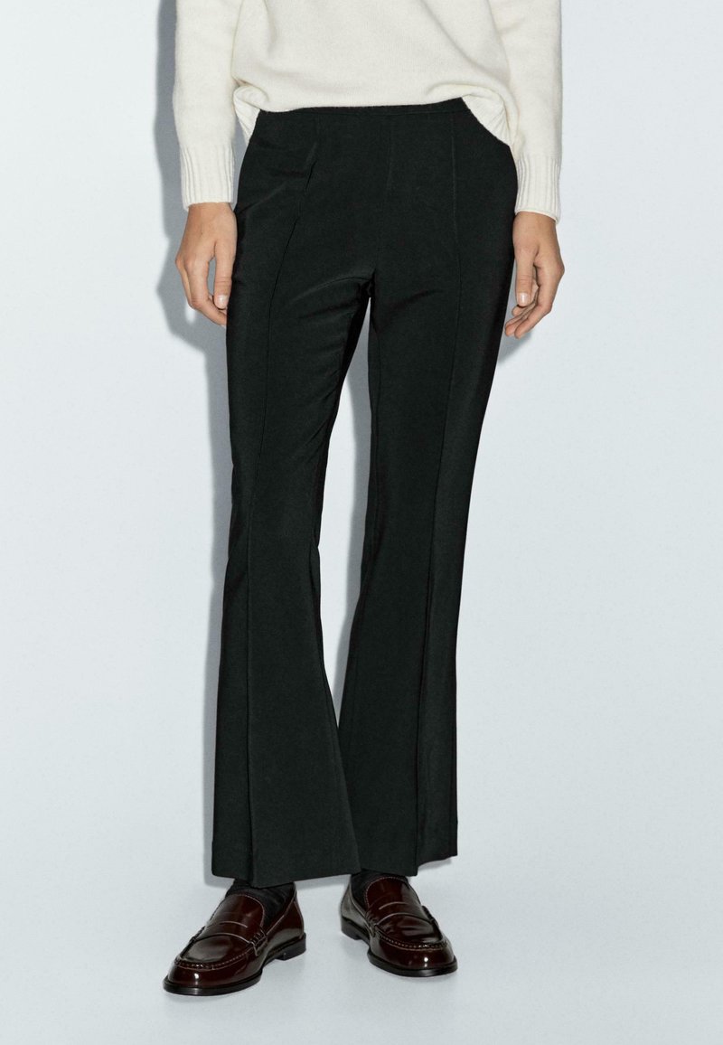 Persona che indossa pantaloni a campana neri su misura, un maglione bianco e mocassini in pelle marrone scuro, in piedi davanti a uno sfondo chiaro e semplice.