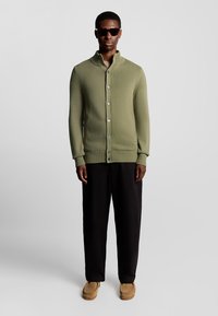 Cardigan en maille vert olive avec un col haut, des fermetures à boutons et des poignets côtelés, associé à un pantalon noir et des chaussures slip-on beiges.