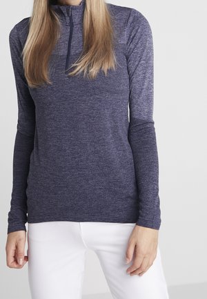 Kobieta ubrana w dopasowany, długorękawowy, ciemnoniebieski sweter z teksturą i suwakiem do ćwiartki, w białych spodniach, stojąca na neutralnym tle.