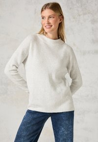 Pull en tricot texturé gris clair avec un col montant et des manches longues, associé à un jean bleu foncé. Design simple, coupe médium.