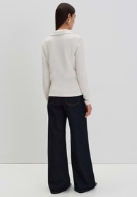 Langärmliger, beiger Pullover mit Kragen, kombiniert mit weit geschnittenen, dunklen Jeans. Weiche Textur und körpernahe Silhouette.