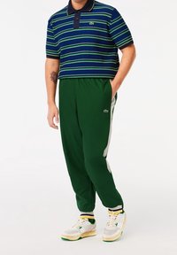 Polo de rayas azul marino y verde con cuello, combinado con pantalones deportivos verdes con detalles laterales de malla. Zapatillas blancas con detalles en verde y amarillo.