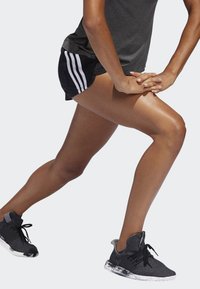 Čierne športové šortky s bielymi pruhmi, šedý športový top a čierne tenisky s textúrovanou tkaninou a vzorovanou podrážkou. Zachytené v pozícii výpadu.