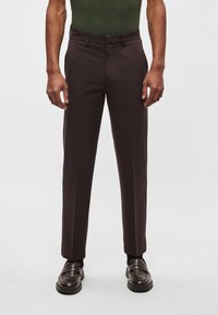 Calças de ganga castanhas feitas de tecido suave, com corte ajustado, frente lisa e bolsos laterais discretos. Usadas com mocassins castanho-escuros.