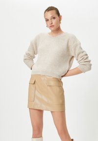 Beżowy sweter o prążkowanej strukturze w luźnym kroju, w zestawieniu z błyszcząco-brązową spódnicą z ekologicznej skóry, wyposażoną w przednie kieszenie. Gładka tekstura i nowoczesne sylwetki.