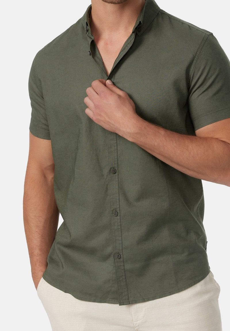 Homme portant une chemise à manches courtes olive à boutons, main gauche ajustant le col, pantalon beige, torse et bras visibles.