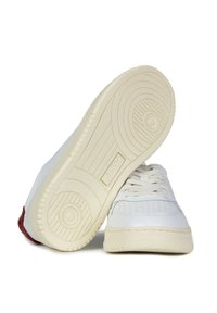 Sneaker bianche in pelle con suola cremoso testurizzata, dotate di perforazioni sulla punta, lacci e un accento rosso sul tallone.