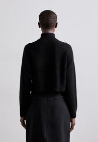 Pull noir court en maille avec un col haut et une texture côtelée, associé à une jupe noire ajustée avec des coutures verticales.
