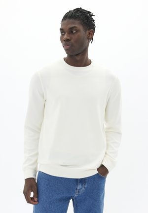 Crème-kleurige sweatshirt met lange mouwen, een ronde hals, aangesloten manchetten en een zachte textuur, gecombineerd met blauwe spijkerbroeken.
