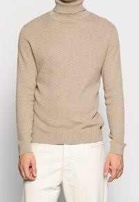 Suéter de cuello alto tejido en beige con un patrón texturizado, cuello y puños acanalados, y corte ajustado. Combinado con pantalones de color claro.