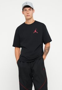 Fekete pamut póló rövid ujjakkal, piros ikonikus Jumpman logóval a mellrészen, laza fekete nadrággal párosítva, piros részletekkel.
