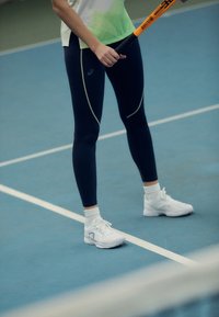 Atletische legging in marineblauw met lichte groene accenten, gecombineerd met witte sportschoenen en sokken, op een blauwe tennisbaan. Houdt een tennisracket vast.