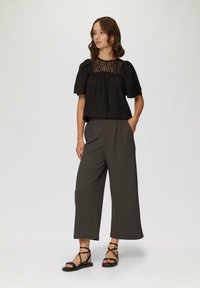 Zwarte top met kantdetails bij de hals en pofmouwen, gecombineerd met donkergrijze, wijduitlopende broeken en zwarte sandalen met bandjes.