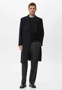 Abrigo de lana azul marino de doble botonadura con suéter negro texturizado y pantalones grises. Zapatos negros completan el conjunto. Diseño limpio y ajustado.
