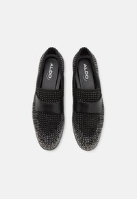Chaussures noires sans lacets avec une bride noire lisse, texture cloutée et bout arrondi. L'intérieur présente une empreinte de logo sur la doublure de la semelle intérieure.