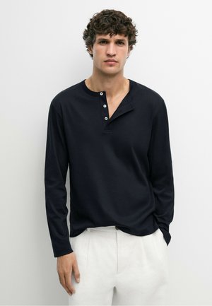Giovane uomo con capelli ricci che indossa una maglia henley nera a maniche lunghe e pantaloni bianchi, in piedi davanti a uno sfondo chiaro uniforme.