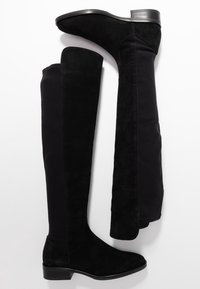 Botas negras por encima de la rodilla con una combinación de materiales de ante y elástico, con un tacón bajo y textura suave, y con un hardware mínimo.