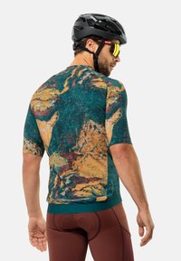 Kurzarm-Fahrradtrikot aus strukturiertem Stoff mit mehrfarbigem abstraktem Muster in Grün-, Orange- und Burgundertönen, kombiniert mit braunen Shorts.