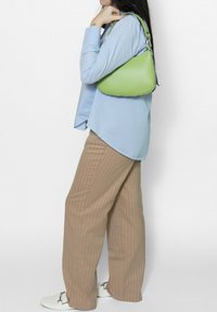 Groene leren schoudertas met gebogen silhouet, gestructureerde afwerking en zilveren hardware, gedragen bij een lichtblauw shirt en gestreepte beige broek.