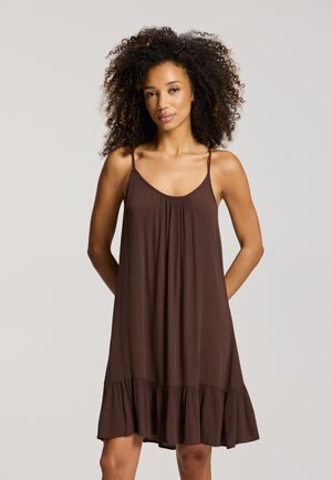 Žena s kučeravými vlasmi, oblečená v hnedých voľných šatách bez rukávov so volánikmi na spodnom okraji, stojaca s rukami za chrbtom.