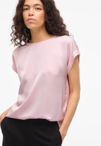 Femme aux cheveux bouclés et foncés, portant un chemisier en satin rose clair à manches courtes et un pantalon noir, les mains dans les poches, sur fond blanc.