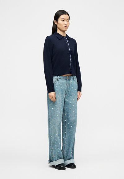 Γυναίκα που στέκεται φορώντας μπλε σκούρο πλεκτό ζακέτα με κουμπιά, ανοιχτό μπλε παντελόνι με φαρδιά μ πατζάκια και μαύρα παπούτσια, μπροστά σε λευκό φόντο.