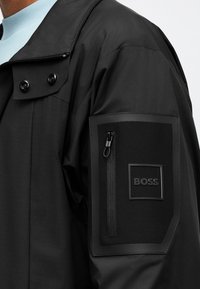 Κοντινό πλάνο μανικιού μαύρου μπουφάν με τσεπάκι με φερμουάρ και λαστιχένιο έμβλημα που απεικονίζει τη λέξη BOSS.