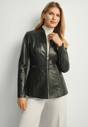 Kobieta o długich blond włosach, ubrana w czarną skórzaną kurtkę na zamek błyskawiczny, założoną na biały golf, oraz jasne spodnie, pozująca na gładkim tle.