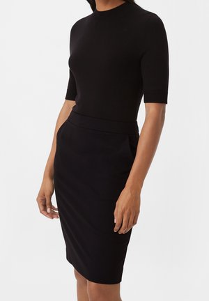 Mujer con una camiseta negra ajustada de manga corta y una falda negra de cintura alta hasta la rodilla, de pie contra un fondo liso.