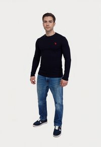 Tmavomodrá košeľa s dlhým rukávom a malým červeným logom, v kombinácii so svetlomodrými džínsami a tmavomodrými teniskami s bielymi akcentmi. Jednoduchý, priliehavý dizajn.