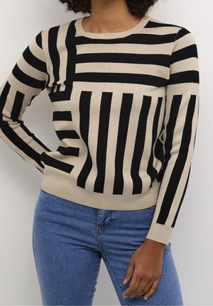 Vrouw in een beige trui met gedurfde zwarte verticale en horizontale strepen, gecombineerd met blauwe spijkerbroeken, staande tegen een eenvoudige witte achtergrond.