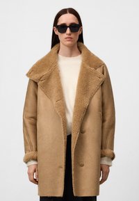 Vrouw in een oversized tan schapenvachtjas met een grote kraag, witte trui, zwarte zonnebril en zwarte broek tegen een effen achtergrond.