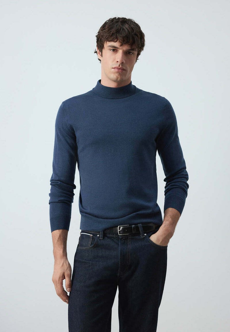 Homme aux cheveux bouclés foncés portant un pull col roulé bleu marine ajusté et un jean foncé, debout avec une main dans la poche.