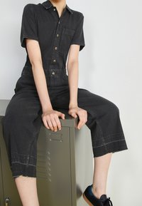 Svart denimoverall med korta ärmar, knappfram, och bröstficka; croppade ben med fransad kant; tillsammans med svarta sneakers.