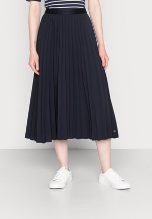 Mujer vestida con una falda midi plisada azul marino con una cintura negra y zapatillas blancas, de pie contra un fondo claro y sencillo.