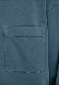 Mörkt teal tyg med en strukturerad yta, med en bröstficka med rundade kanter och synliga sömnadsdetaljer.