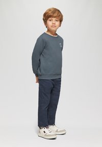Jongentje met lichtbruin haar staat met zijn gezicht naar voren, gekleed in een donkerblauwe sweatshirt, marineblauwe broek en witte sneakers tegen een effen achtergrond.