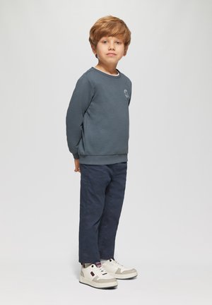 Jongentje met lichtbruin haar staat met zijn gezicht naar voren, gekleed in een donkerblauwe sweatshirt, marineblauwe broek en witte sneakers tegen een effen achtergrond.