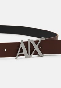 Brauner Ledergürtel mit einem eleganten schwarzen Innenfutter, ausgestattet mit einer auffälligen silbernen Logo-Schließe mit stilisierten Buchstaben "AIX."