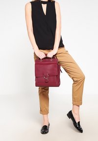 Zaino in pelle bordeaux con patta frontale e accessori decorativi, abbinato a pantaloni beige e un top nero senza maniche. Mocassini neri.