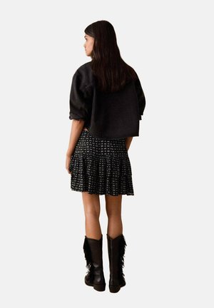 Veste noire surdimensionnée, jupe à volants noire à motif avec des détails floraux, et bottes noires montantes avec des accents à franges. Vue de dos.