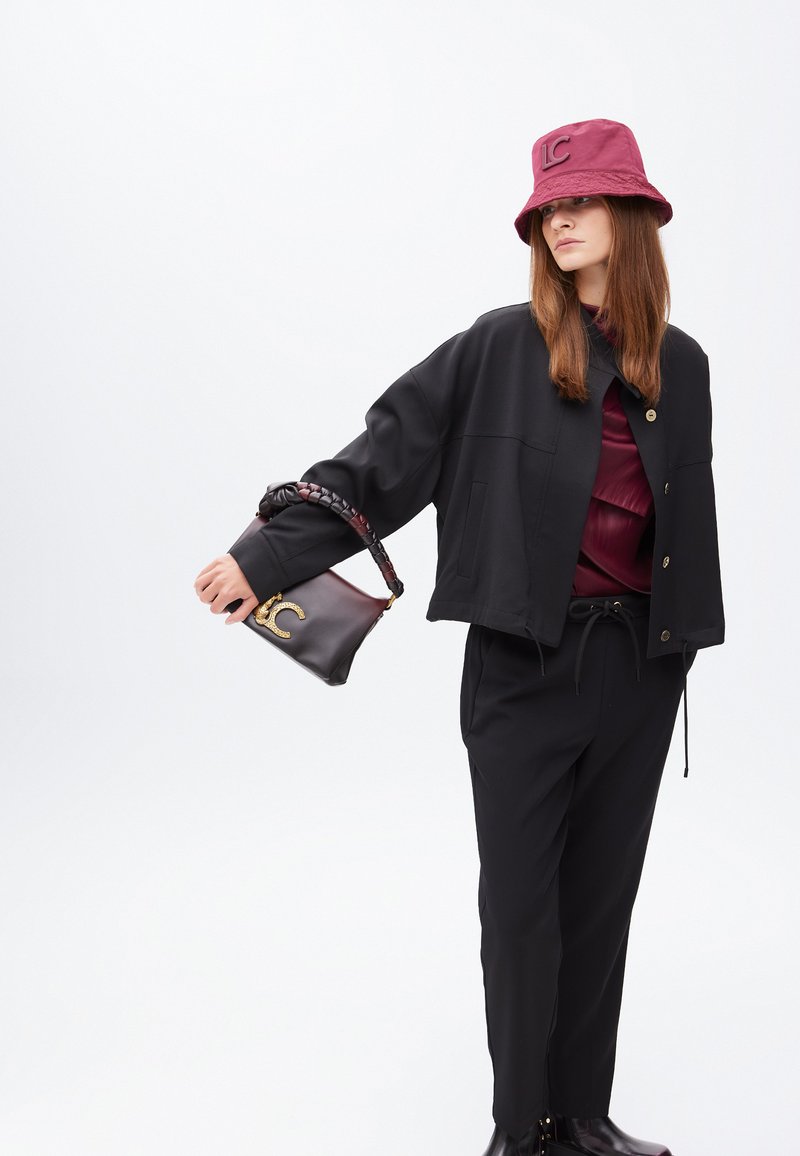 Una modella posa indossando una giacca nera, un top bordeaux, pantaloni neri e un cappello a secchiello abbinato, mentre tiene una borsa con un logo.