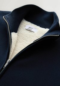Veste marine zippée avec un extérieur texturé et une doublure en polaire douce crème. Comprend un col côtelé et une fermeture éclair en métal. Étiquette de taille visible.