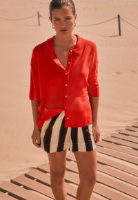 Rotes kurzärmeliges Hemd mit Knöpfen, kombiniert mit schwarz-weiß gestreiften Shorts. Das Hemd hat einen Kragen und Knöpfe und eine lockere Passform.