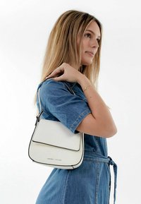 Bolso blanco de hombro con borde negro, de forma rectangular, textura de cuero suave y solapa frontal. Correa ajustable para un uso versátil.