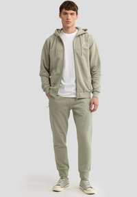 Hellgrüner Hoodie und Jogginghose aus weichem Stoff, mit einem weißen Logo auf der linken Brust und an der Hose, sowie gerippten Bündchen und Kordeln.