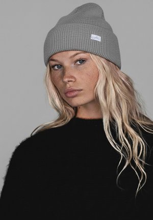 Gray ribbed beanie with a small label, worn atop long, wavy hair. Black sweater with a crew neck visible beneath the beanie.