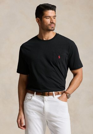 Mann trägt schwarzes kurzärmeliges T-Shirt mit rotem Logo auf der Tasche, weiße Hose, brauner Gürtel und Uhr, vor beigem Hintergrund.