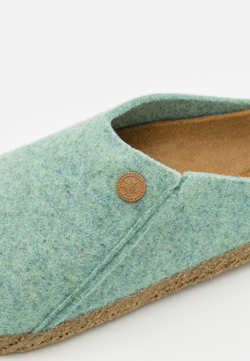 Filz-Schlupfschuh in hellem Blaugrün mit rundem Zehenbereich, Korksohle und einem kreisförmigen Logo-Detail an der Seite. Weicher Textur mit minimalem Nähen.
